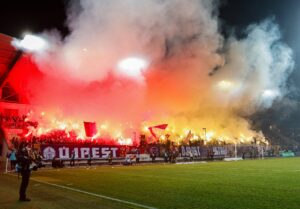 Újpest-szurkolók pirotechnikával és zászlókkal támogatják csapatukat egy mérkőzésen.